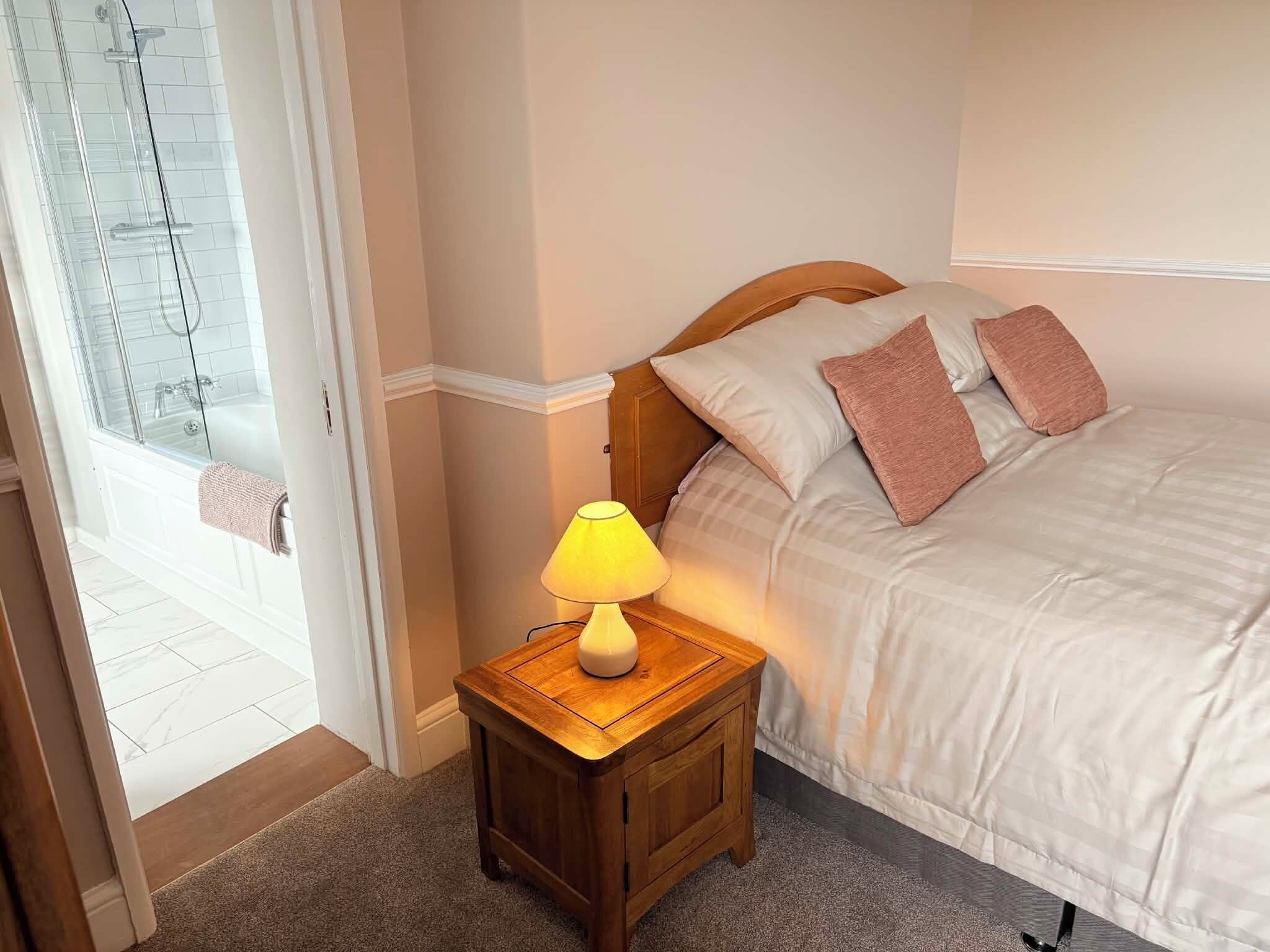 A bedroom with a bed, a bedside lamp, and a view of a bathroom with a glass shower enclosure.