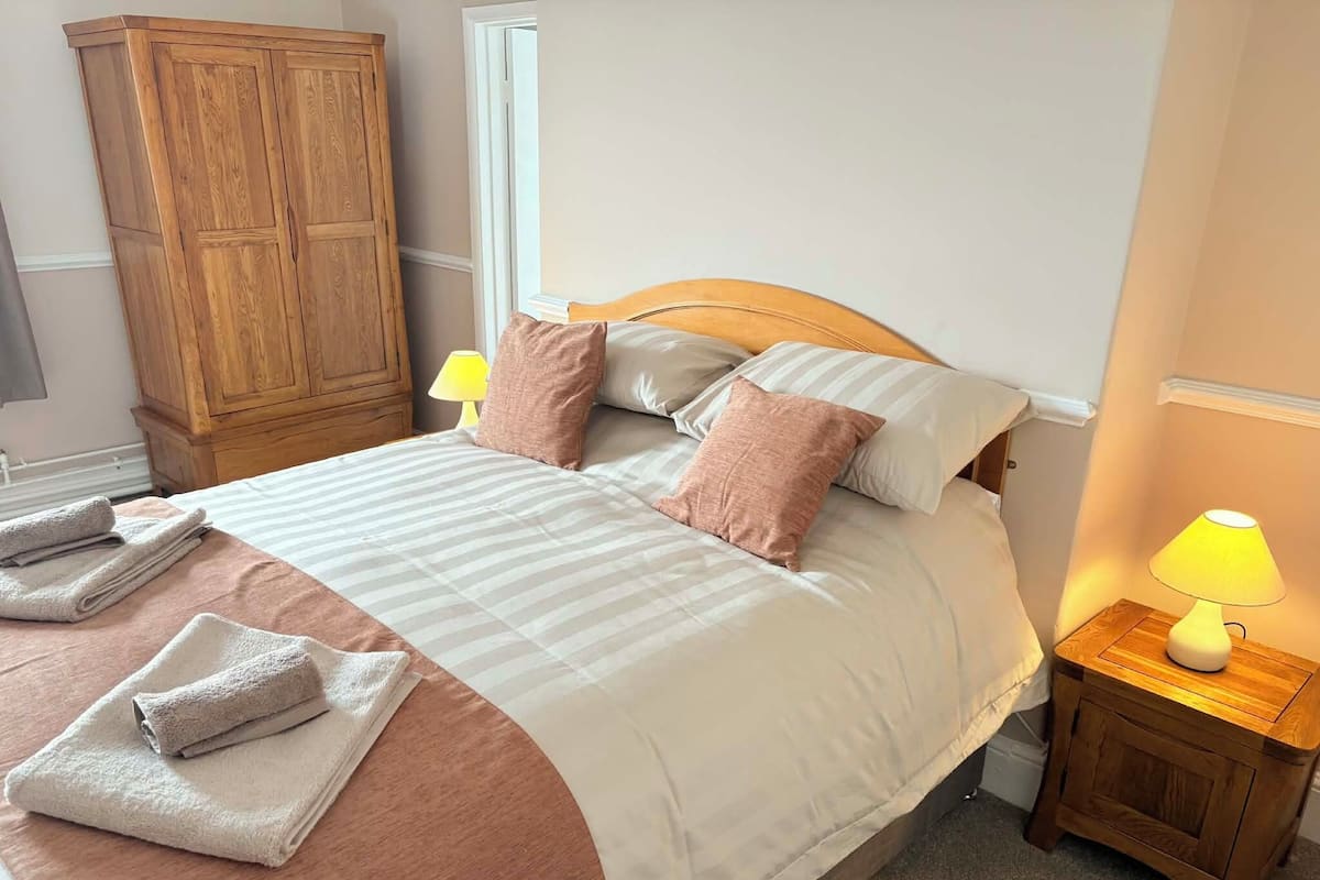 A neatly made bed with pillows, a wooden wardrobe, a bedside lamp, and folded towels.