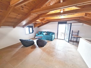 An attic room with a wooden ceiling, a blue sofa, two bean bags, a small round table, and a sliding door leading outside. - Viki's Sea View Apartment (Lefkada)