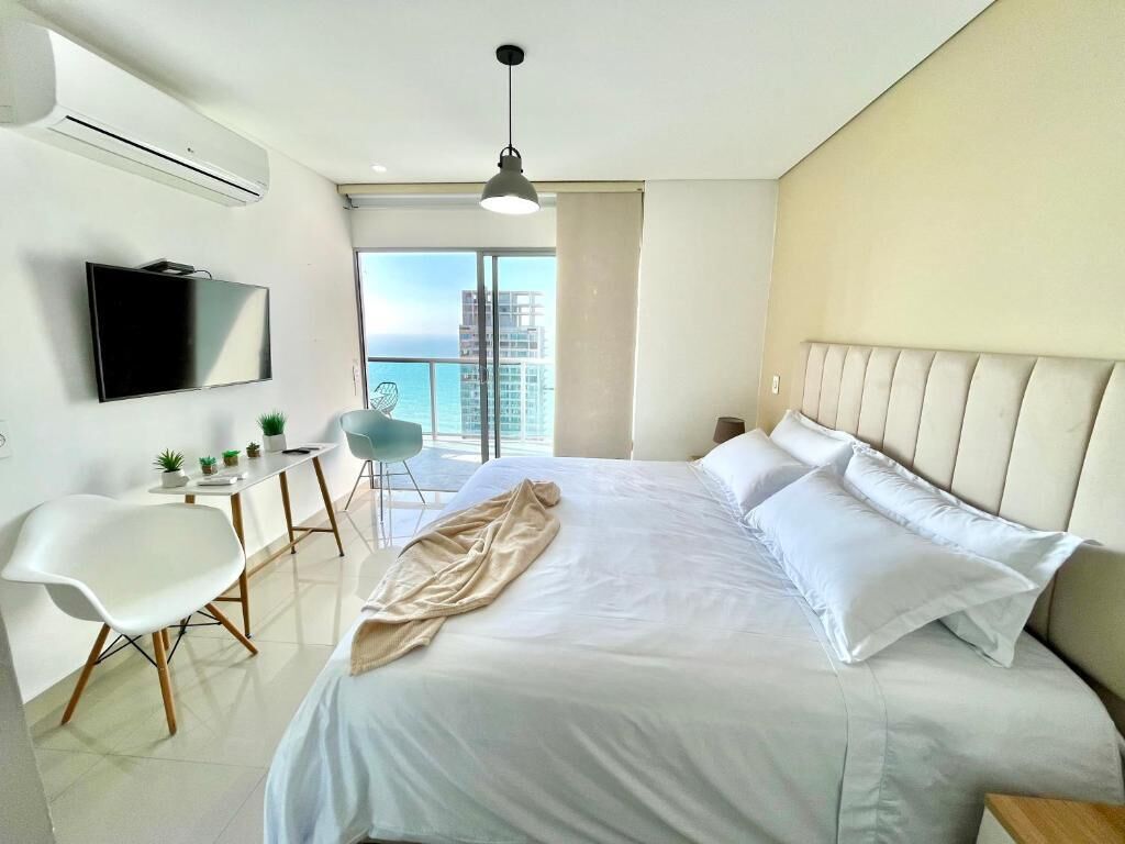 A modern hotel room with a bed, a TV, a balcony with a view, and a dining area.