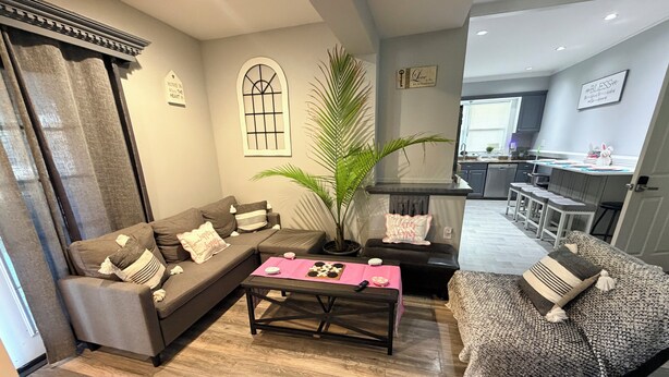 A living room with a sectional sofa, a coffee table, a patterned armchair, a potted plant, and kitchen area in the background. - Cozy 2-bedroom apartment in beautiful Ewing Township with AC (Ewing Township)
