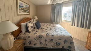 Une chambre à coucher avec un lit, un ventilateur de plafond, une lampe, un tableau et une fenêtre avec des rideaux.