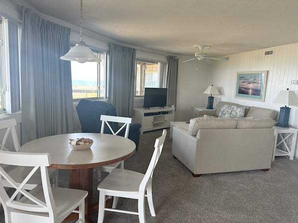 A hotel room with a dining table, chairs, a sofa, a TV, a lamp, and a painting on the wall.