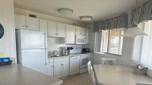 Une cuisine avec des meubles blancs, un réfrigérateur, un four à micro-ondes, une cuisinière et un évier. On y trouve une table à manger avec des chaises et une fenêtre avec des rideaux.