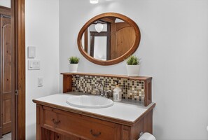 A bathroom with a wooden vanity, a round mirror, a sink, and two potted plants. - Les Manoirs Unit 9 (Mont-Tremblant)