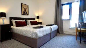 Habitación de hotel con cama, dos mesitas de noche, una silla, ventana con cortinas e imagen colgada en la pared.