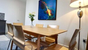 Un comedor con mesa de madera, seis sillas, un florero con flores, un cuadro abstracto colorido en la pared y una lámpara de pie.