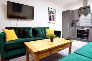A living room with a green sofa, a wooden coffee table, a flat-screen TV, a framed picture, and a kitchen with a microwave and oven. - B - Delux 2-bedroom apartment in the center of Thatcham (Thatcham)