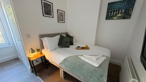 Ein ordentlich bezogenes Bett mit Kissen, ein Nachttisch mit Lampe und Kunstwerke an den Wänden.