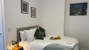 Ein ordentlich bezogenes Bett mit Kissen, ein Nachttisch mit Lampe und eine Ablage mit Früchten und einem Getränk.