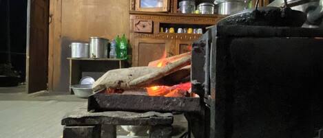 A rustic kitchen with a wood-fired oven, a shelf with pots, and a wooden cabinet.