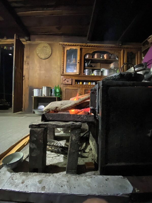 A rustic kitchen with a wood-fired oven, a shelf with pots, and a wooden cabinet.