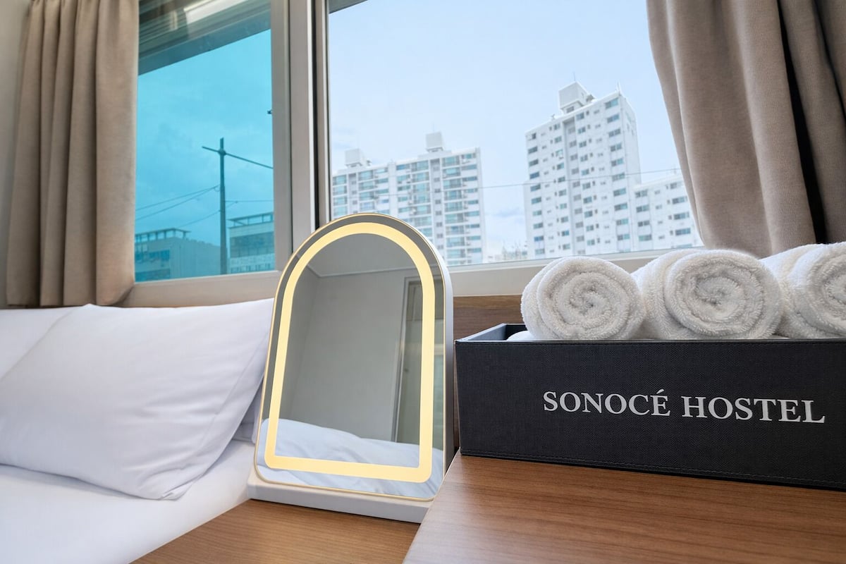 A hotel room with a bed, a bedside table with a mirror, a box of towels, and a window with a city view.