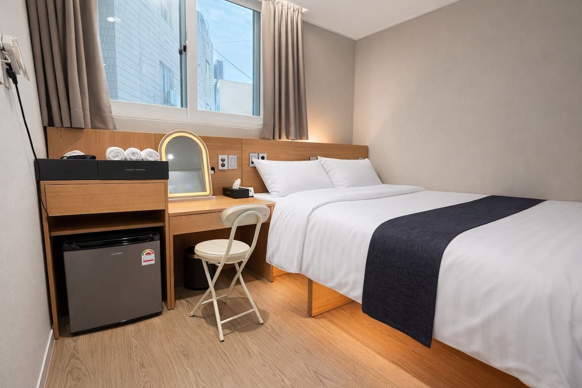 A compact hotel room with a bed, a desk with a mirror, a small refrigerator, and a chair.