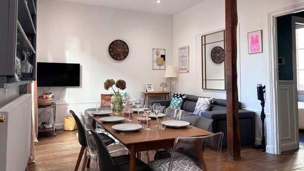 A dining area with a wooden table set for four, surrounded by chairs. There is a television mounted on the wall, a clock, and a framed picture. - Particulier rémois proche Cathédrale avec parking et terrasse (Reims)