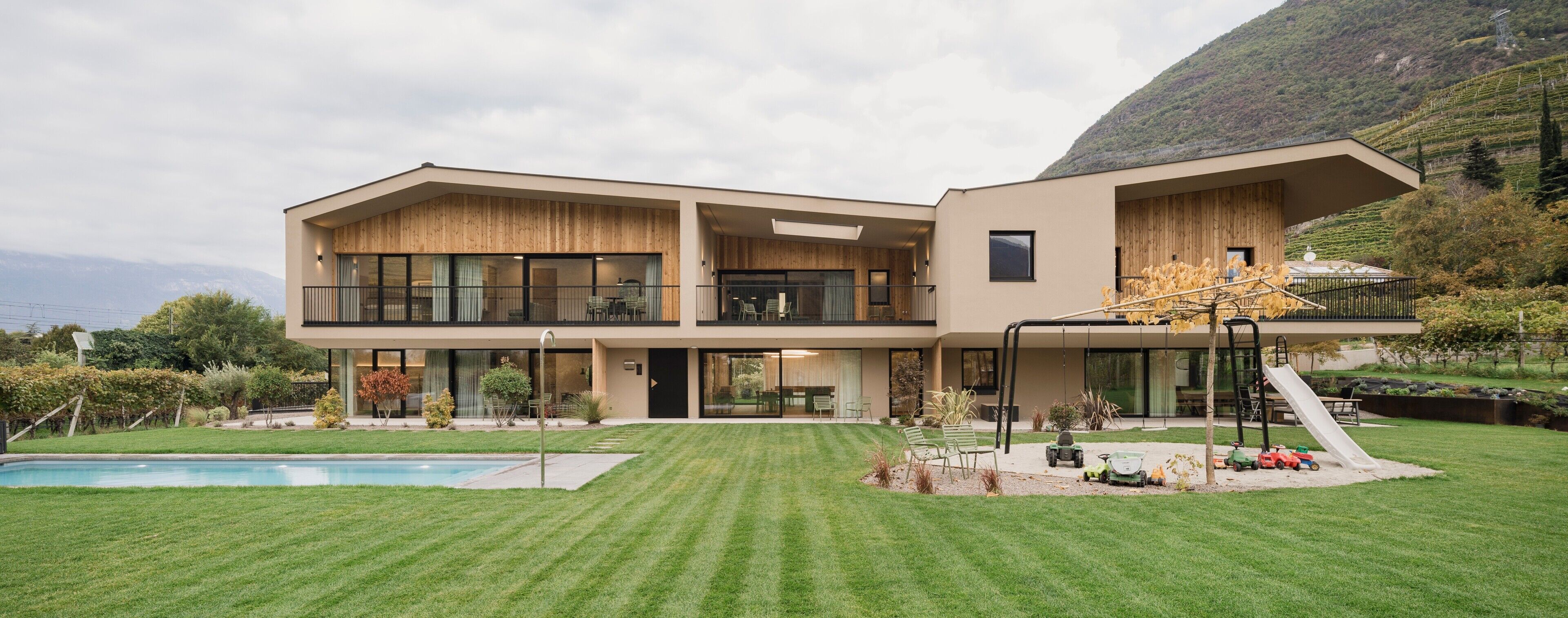 A modern two-story house with a swimming pool, a covered outdoor seating area, and a view of a mountainous landscape.