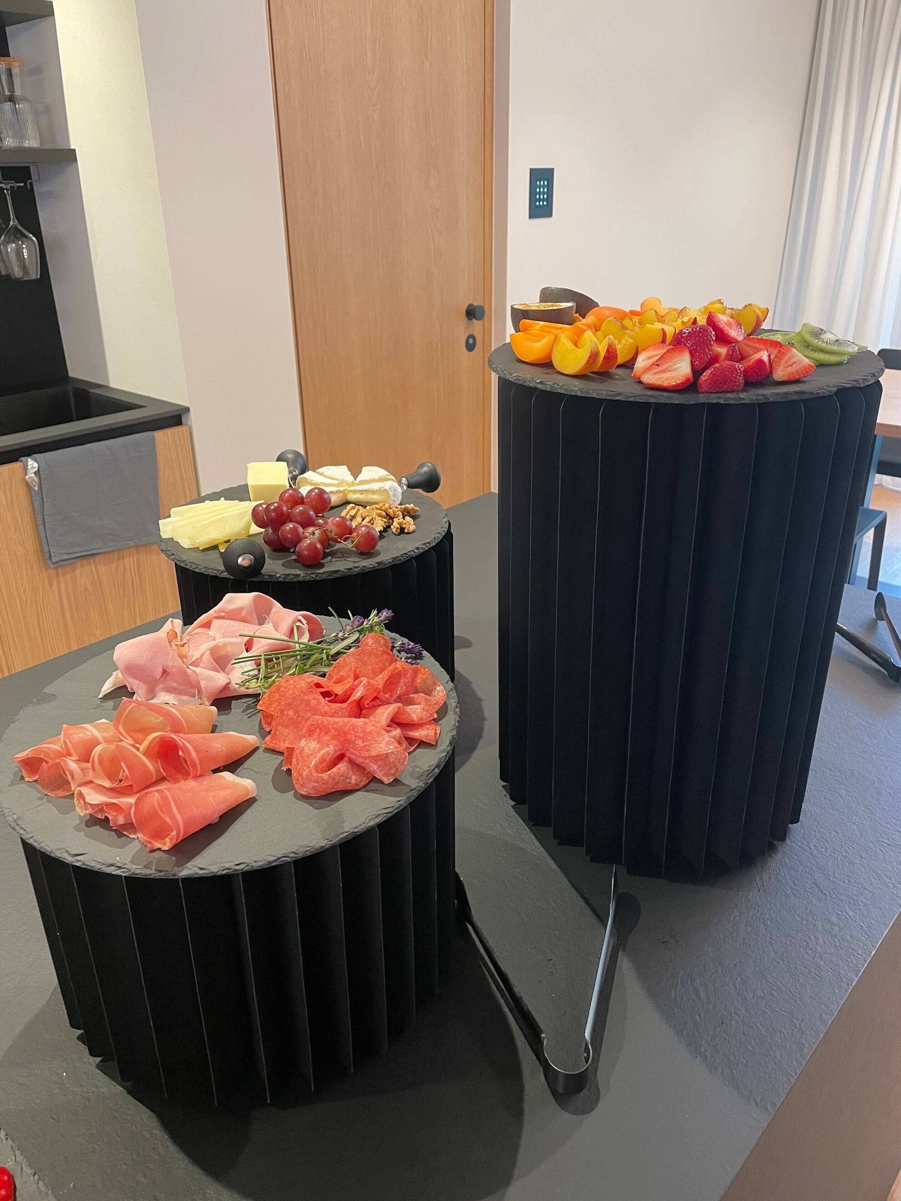 A room with a kitchen area, a dining table with a fruit platter, and a meat platter.