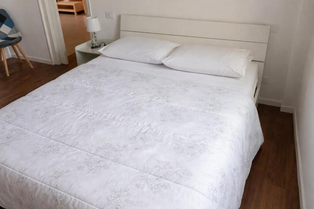 A neatly made bed with a white bedspread, a bedside table with a lamp, and a chair in a room with wooden flooring and white walls.