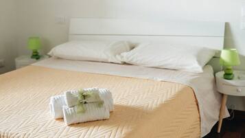 Una cama bien hecha con colcha color beige, dos almohadas y un juego de toallas sobre la cama. Hay dos mesitas de noche, una con lámpara verde y otra con lámpara blanca. La habitación tiene piso de madera y paredes blancas.