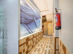 A hallway with a patterned floor, a wall mural, a wooden door labeled 'TOILET,' and a fire extinguisher mounted on the wall. - Deluxe Hotel Shikhar Darshan (Ujjain)
