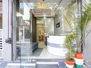 A reception area with a curved counter, a glass door entrance, a ceiling fan, and a potted plant. - Deluxe Hotel Shikhar Darshan (Ujjain)
