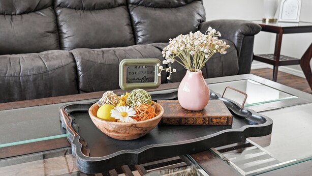 Een moderne woonkamer met een leren bank, een glazen salontafel, een houten schaal met gedroogde bloemen, een roze vaas en een dienblad met een waterkoker.