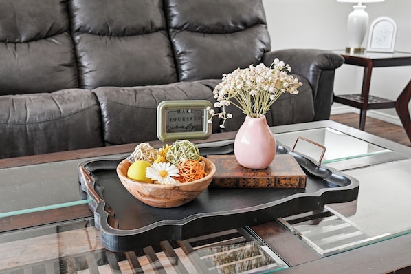 Un salón moderno con un sofá de cuero, una mesa de centro de vidrio, un cuenco de madera con flores secas, un jarrón rosa y una bandeja con un hervidor de agua.
