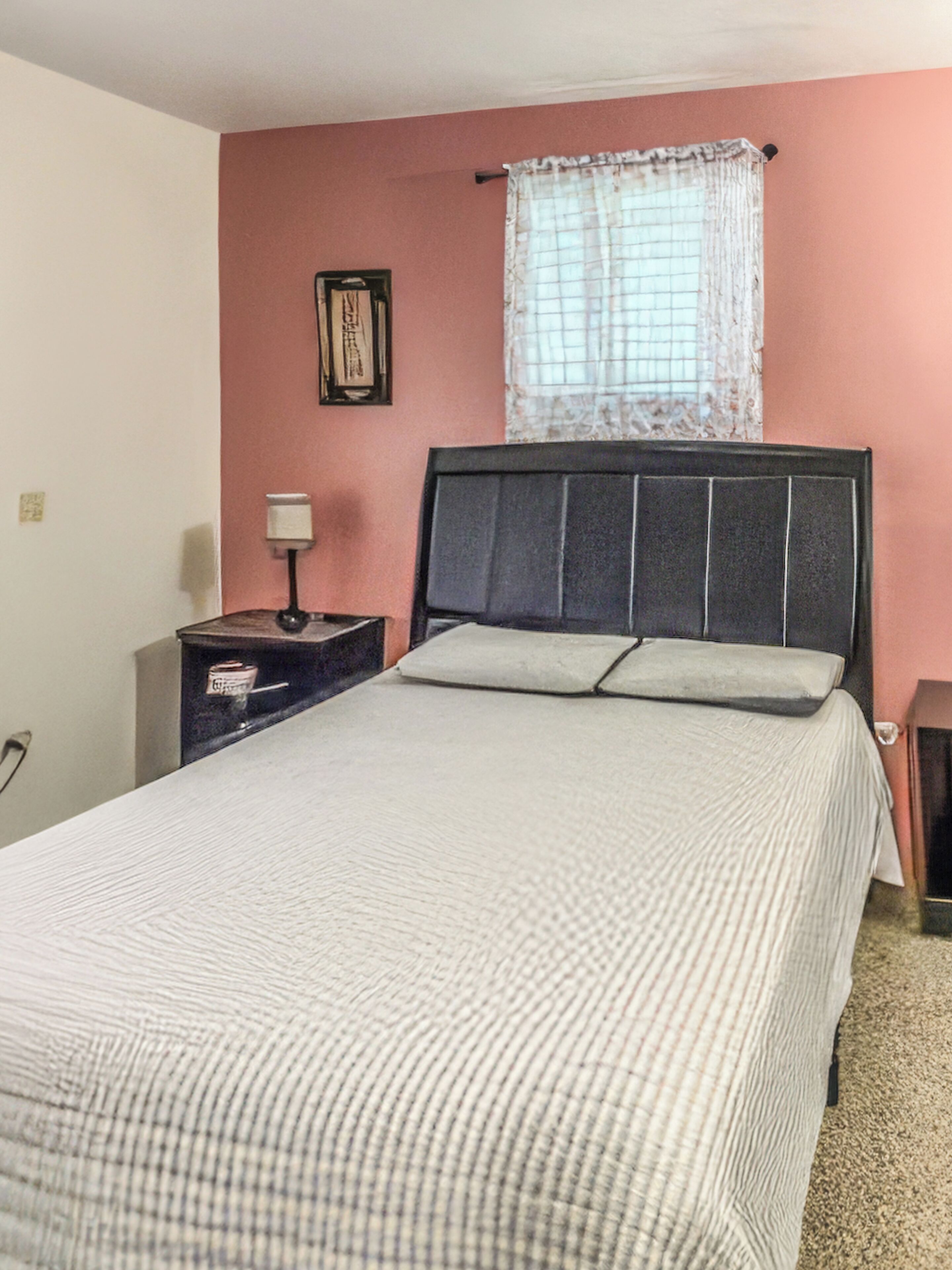 Un dormitorio con cama, mesita de noche, lámpara, ventana con cortina e imagen colgada en la pared.