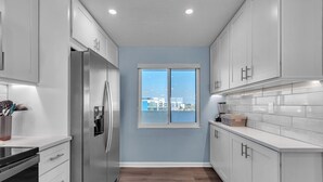 A modern kitchen with white cabinets, a stainless steel refrigerator, a window with a view, and a wooden floor. - Ocean Sands 2-602 (Madeira Beach)
