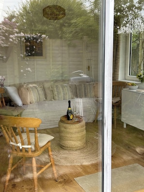A cozy living room with a sofa, a wooden chair, a round coffee table with a bottle and glasses, and a view of greenery outside.