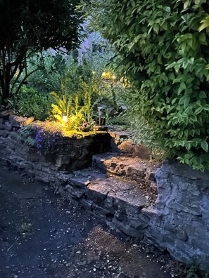 A stone pathway leading through a garden with various plants and a small water feature.