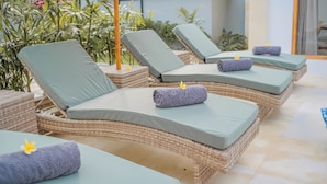 A row of wicker lounge chairs with cushions, arranged outdoors with a view of greenery and a building in the background. - Dolphin 2 – Deluxe Double Room in New Villa on Lovina Beach (Buleleng)