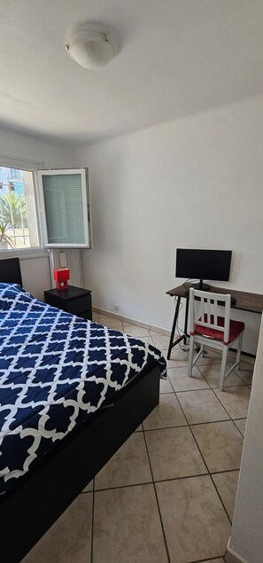 Une chambre à coucher avec un lit, un bureau avec une chaise, une fenêtre avec des stores et un radiateur.