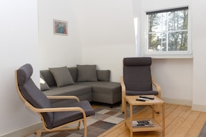 Un salon comprenant un canapé d’angle, deux fauteuils, une petite table en bois et une fenêtre donnant sur des arbres.
