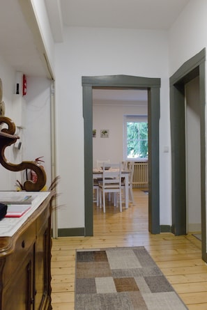 A hallway with a wooden floor, a rug, a dining area with white chairs and a table, and a door leading to another room.