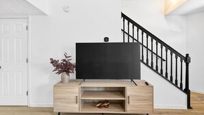 Ein modernes Wohnzimmer mit Flachbildfernseher auf einer Holzkonsole, einer Treppe mit schwarzen Geländern und einer weißen Tür.
