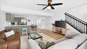 Ein modernes Wohnzimmer mit einem braunen Ledersofa, einem grauen Sofa, einem Glastisch, einem Fernseher auf einem Holzständer und einem Deckenventilator.
