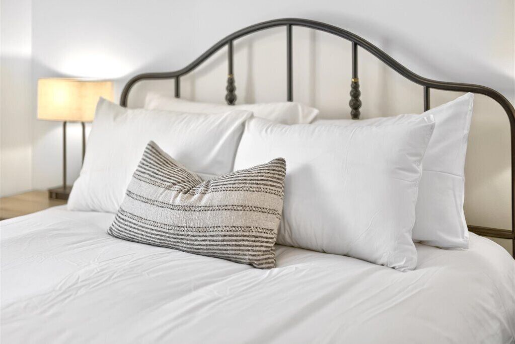 Ein Bett mit weißer Bettwäsche und zwei Kissen, eines gestreift, eines einfarbig, in einem Zimmer mit einem dunklen Metall-Kopfteil und einer Nachttischlampe.