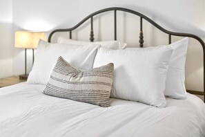Una cama con ropa de cama blanca y dos almohadas, una a rayas y otra lisa, en una habitación con cabecera metálica oscura y una lámpara de noche.