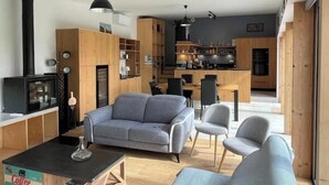 Ett modernt vardagsrum med en soffa, fåtöljer, ett soffbord och en tv. Det finns också ett köksområde med skåp och ett matbord.