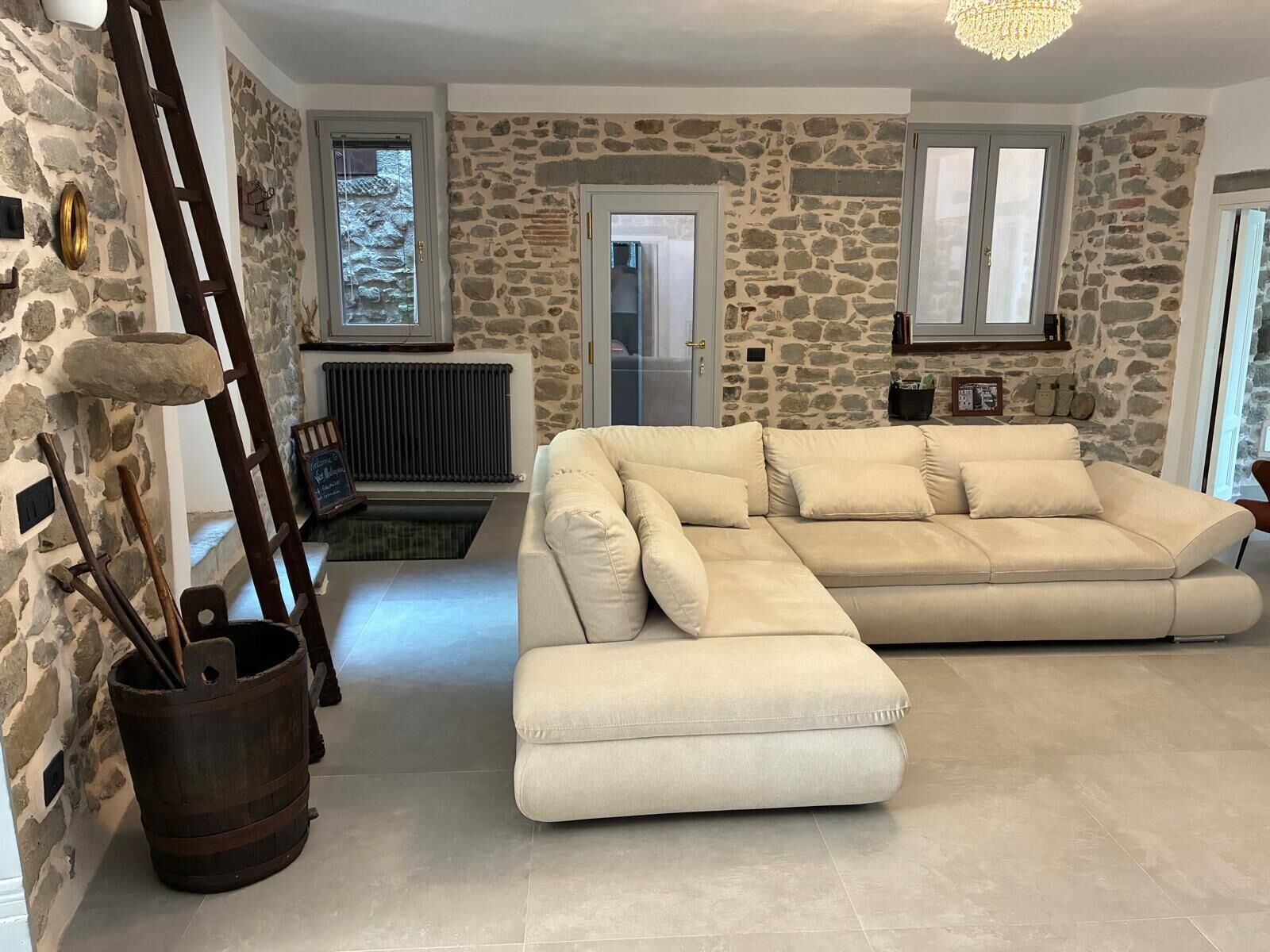 Un salon avec un mur en pierre, un grand canapé d’angle beige, une échelle en bois et un radiateur.