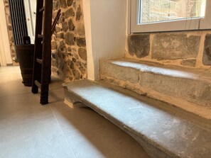 Una escalera de piedra que sube a un nivel superior, con una silla de madera y un muro de piedra al fondo.