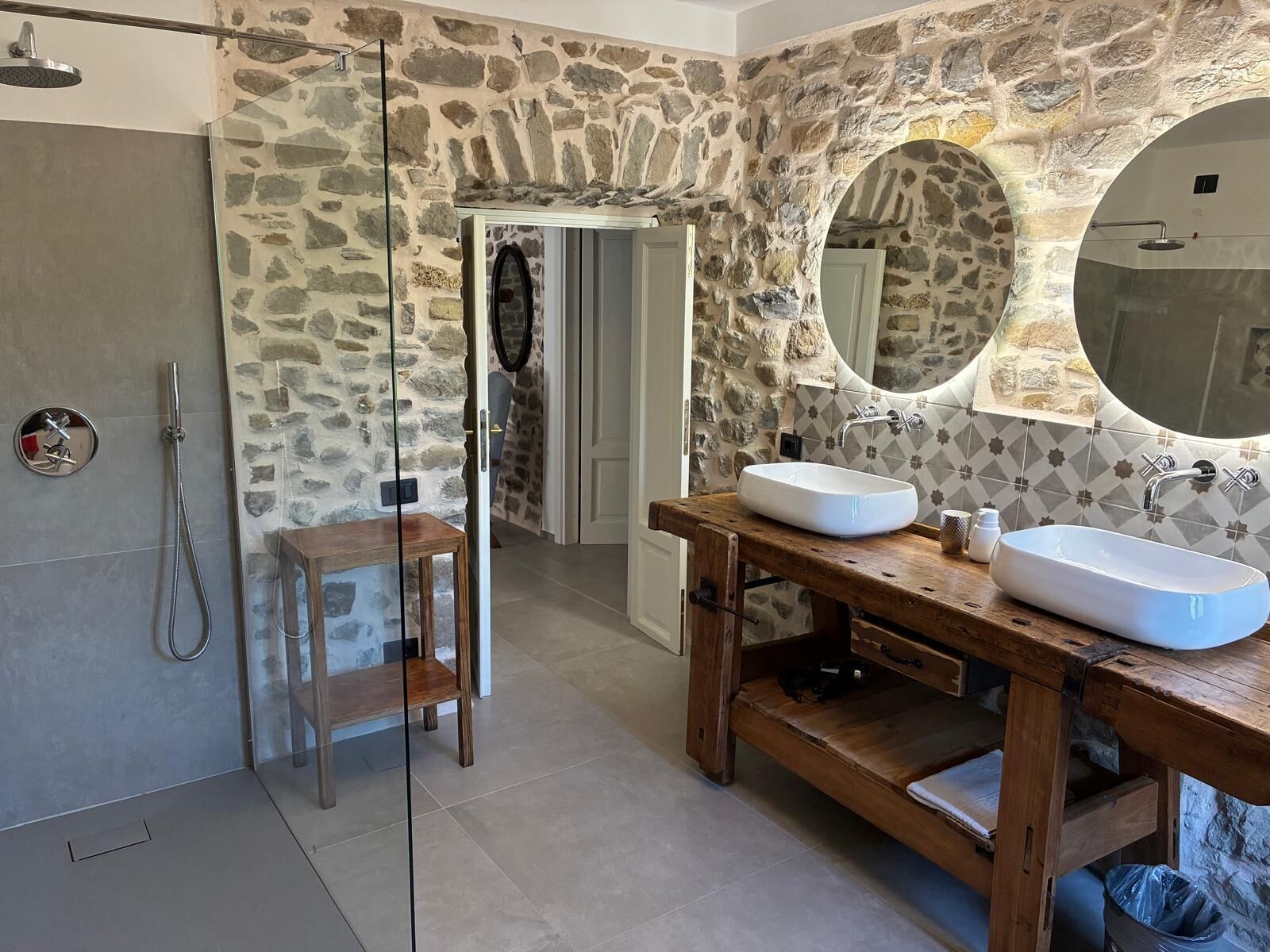Une salle de bain avec un mur en pierre, un miroir rond, deux lavabos et une douche.