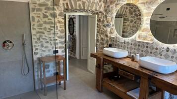 Une salle de bain avec un mur en pierre, un miroir rond, deux lavabos et une douche.
