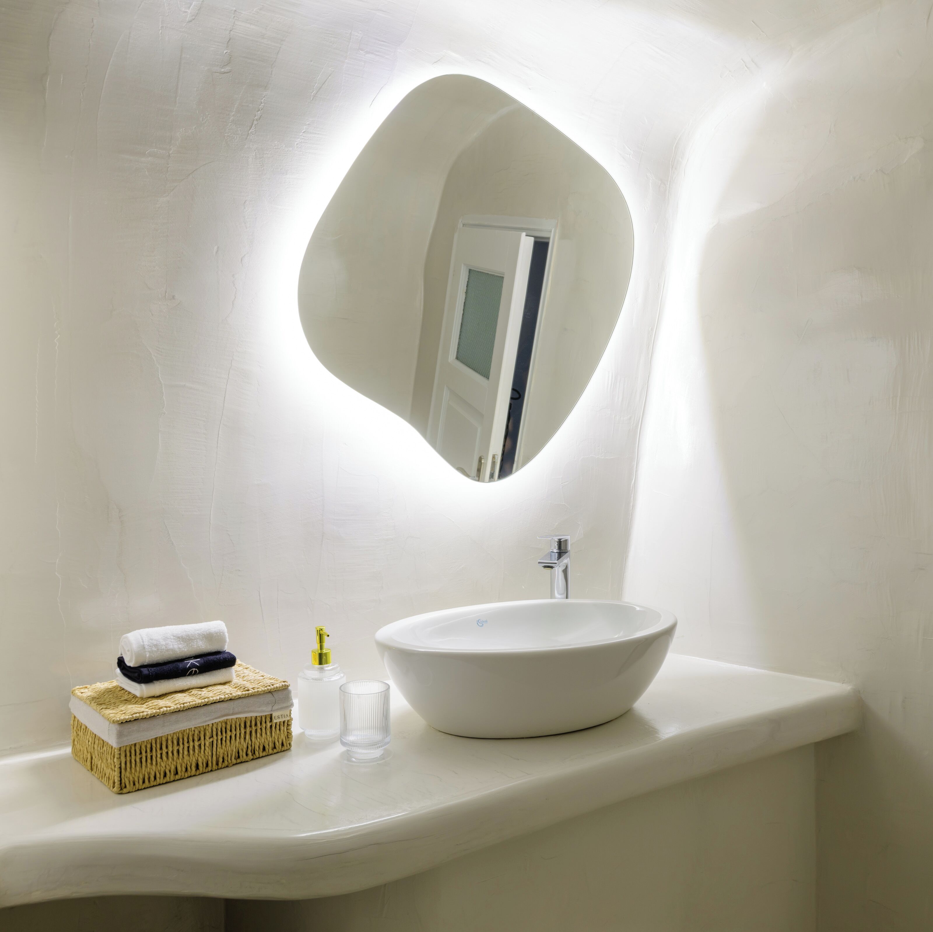 Un baño con un lavamanos blanco redondo, un espejo de forma original y una pequeña ventana.