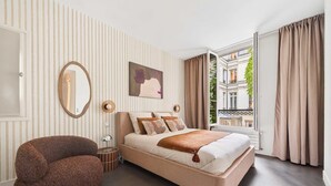 Un dormitorio con cama, una silla, ventana con cortinas, espejo y un cuadro en la pared.