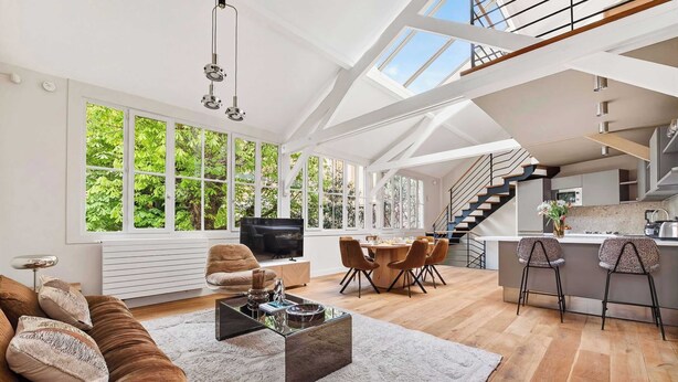 Amplia sala de estar con techos altos, ventanas grandes, zona de comedor y cocina con una escalera.