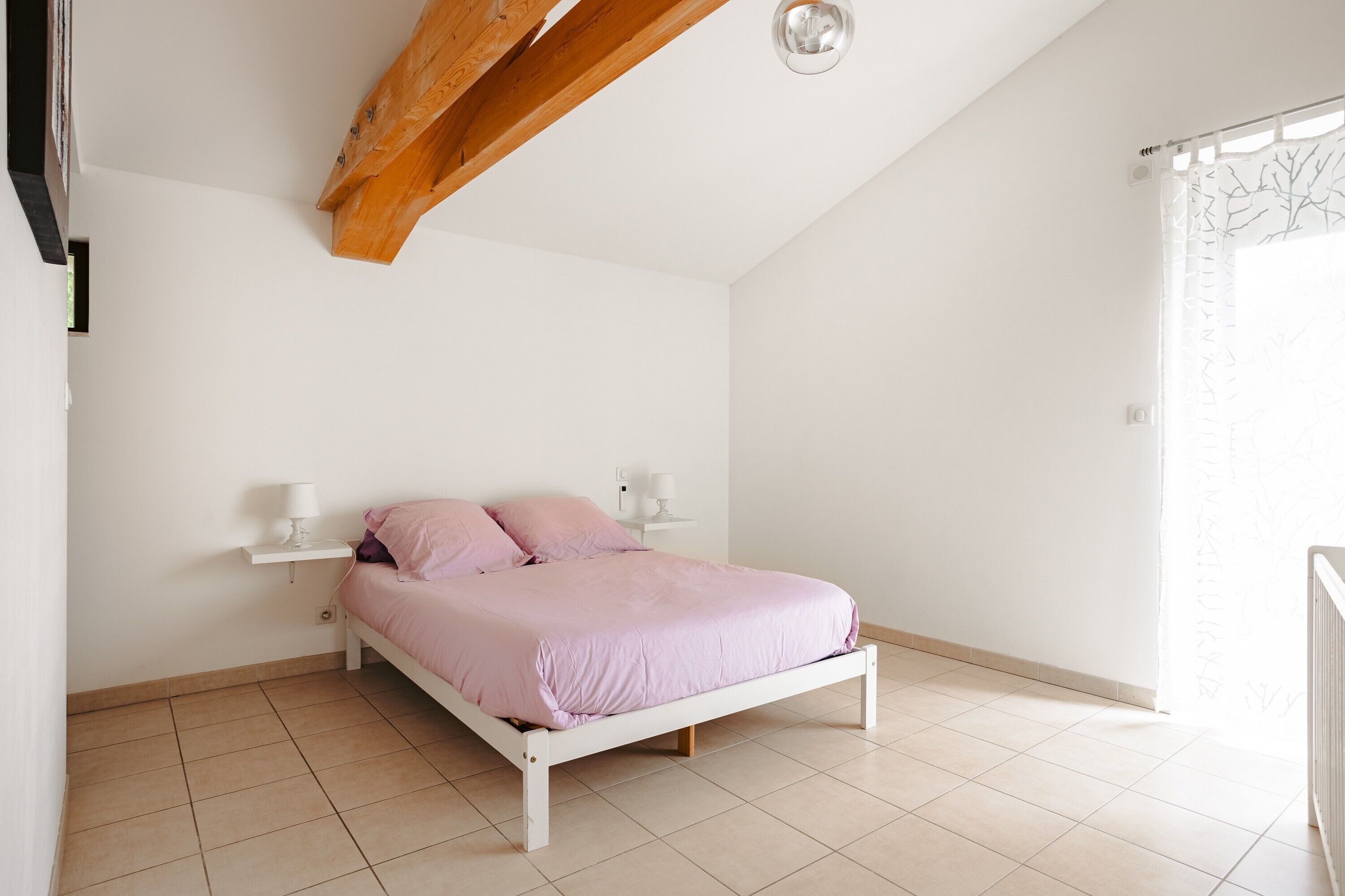 A bedroom with a bed, bedside lamps, a wooden ceiling beam, and a window with a curtain.
