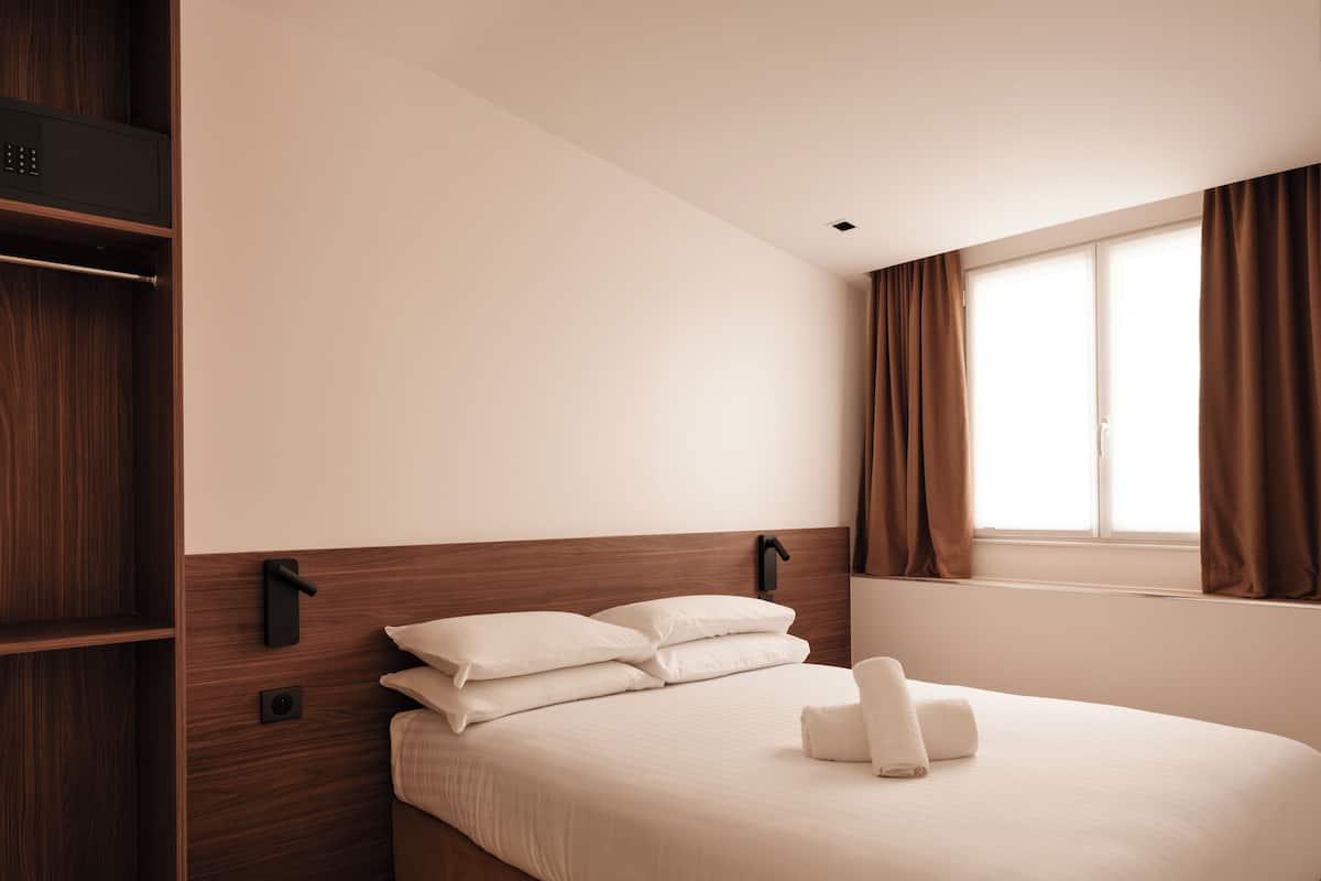 Ein Hotelzimmer mit Holzkopfende, einem Bett mit weißer Bettwäsche, einem Nachttisch und einem Fenster mit Vorhängen.