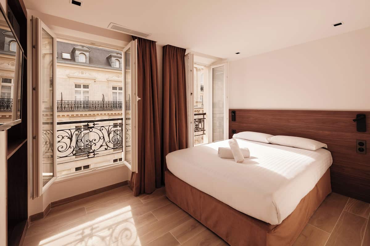 Ein Hotelzimmer mit Bett, Fenster mit Vorhängen, Balkon mit Geländer und einem hölzernen Kopfteil.